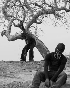 A black-and-white photograph shows two masked figures near a gnarled tree on barren ground, one standing and one seated.