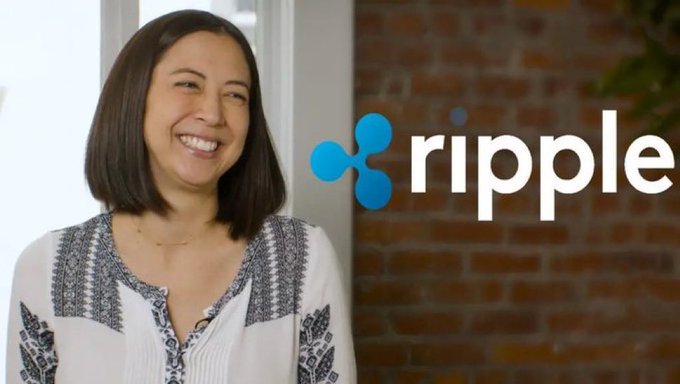 A smiling woman with shoulder-length brown hair standing against a brick wall background with the Ripple logo displayed beside her.