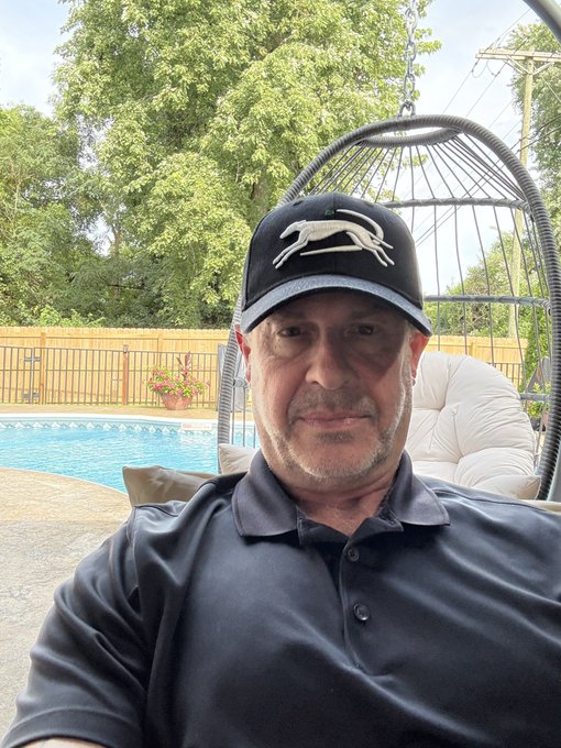 A selfie of a man wearing a black cap with a greyhound logo and a black polo shirt, taken outdoors near a swimming pool with a hanging chair and wooden fence in the background.