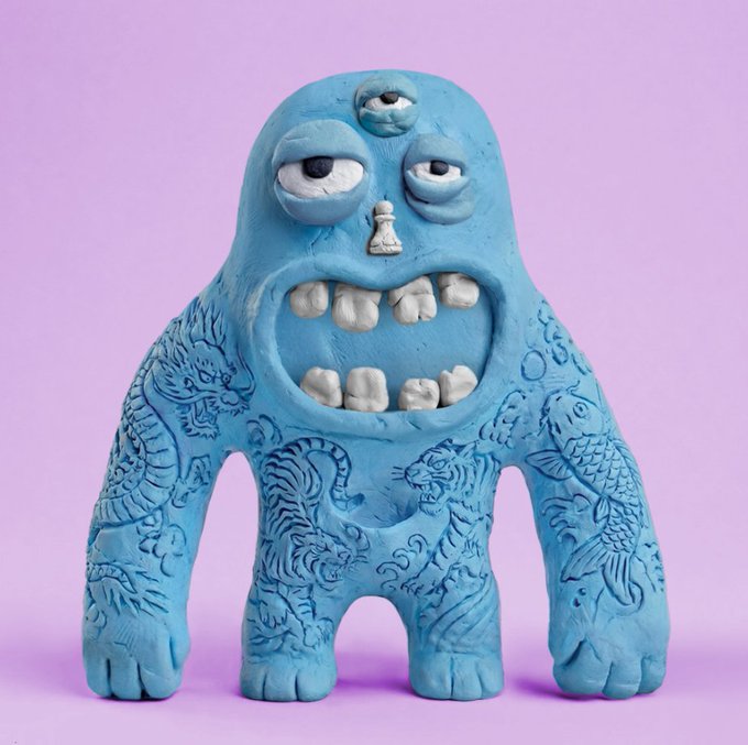 A blue clay sculpture of a quirky three-eyed monster with a wide toothy grin, covered in intricate animal and floral etchings, photographed against a pink background.