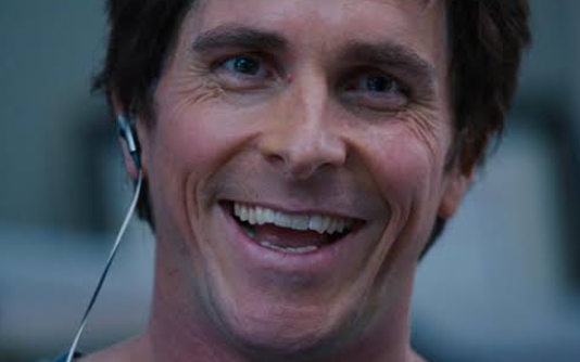 A close-up photo of a smiling man wearing earbuds with white cables, resembling a celebrity or public figure.