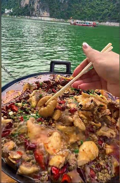 A hand uses chopsticks to pick up food from a large pan of spicy stir-fry, with a green body of water and a boat visible in the background.