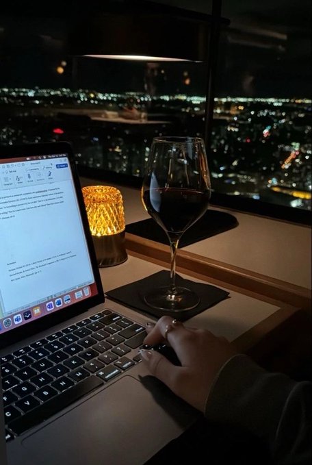 The image shows a person sitting at a desk with an open laptop computer, typing on its keyboard while enjoying a glass of wine. There are two candles on the desk, one near the left side and another closer to the right edge of the scene. The background features a city skyline, suggesting that they might be in a restaurant or a similar urban setting.