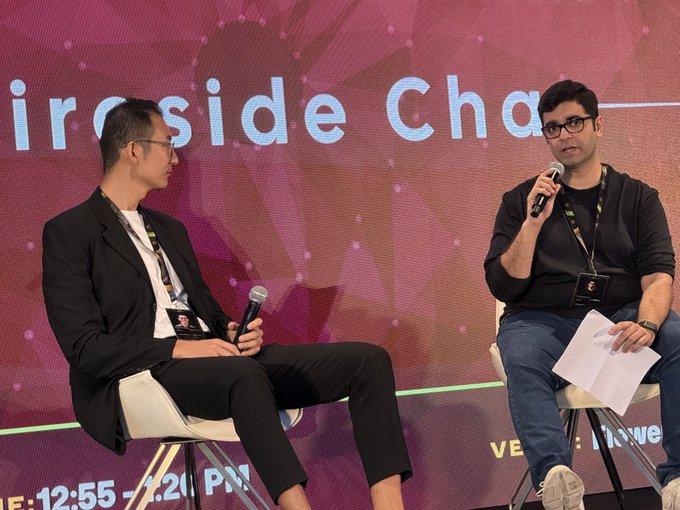 Two men sitting on stools holding microphones during a 'Fireside Chat' panel discussion, with event branding in the background.