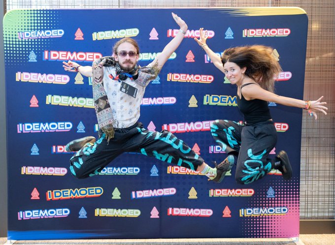 Two people joyfully jump in the air in front of a colorful backdrop covered with 'IDEMOED' logos and graphics.