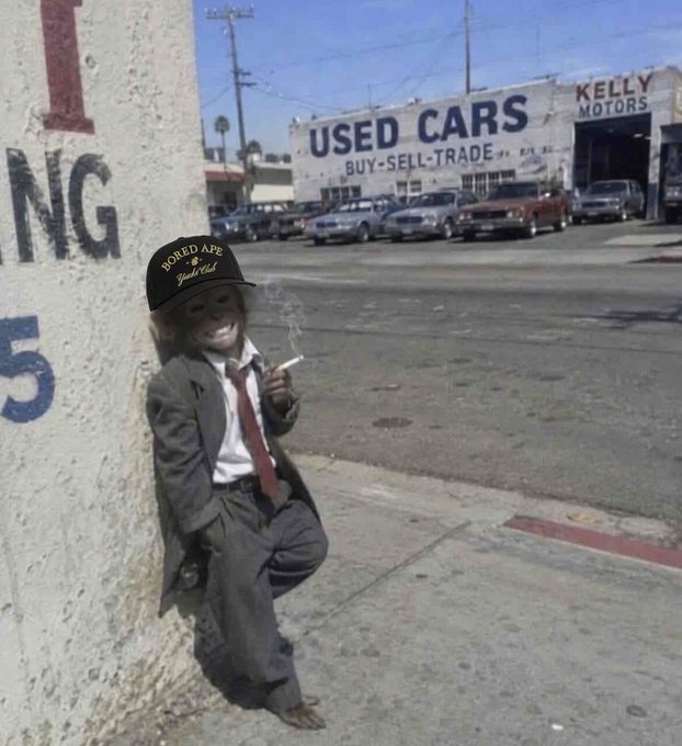 The image features a young boy wearing a suit, tie, and baseball cap with the words "Bored Ape" written on it. He is leaning against a wall next to a car dealership, holding a cigarette. The caption of the image reads, "A Bored Ape is a way of life." This suggests that the boy's attire and actions are representative of the post's message about the importance of finding joy in everyday life.