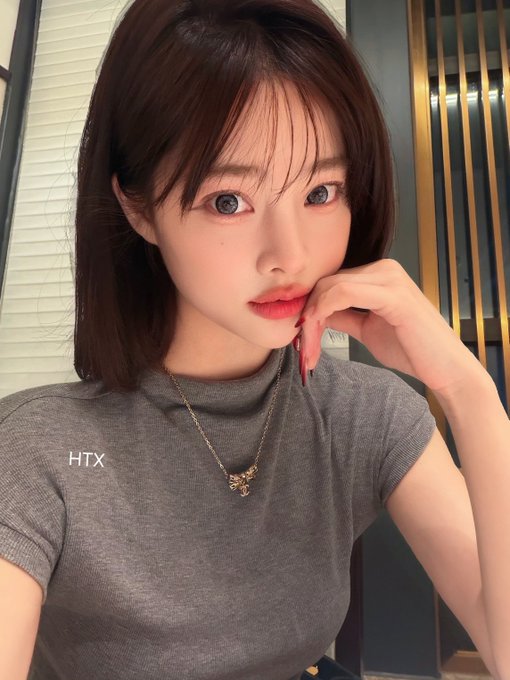 A woman with long brown hair and blue-gray eyes poses with her hand near her face, wearing a fitted gray t-shirt with "HTX" printed on it and a gold pendant necklace. The "HTX" branding on her shirt ties into the post's promotion of HTX Prime VIP membership benefits, with her polished appearance complementing the premium service message.