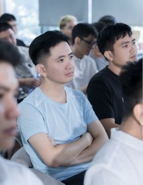 A young man in a light blue t-shirt sits with his arms crossed, attentively watching something in what appears to be a classroom or presentation setting, surrounded by other attendees.
