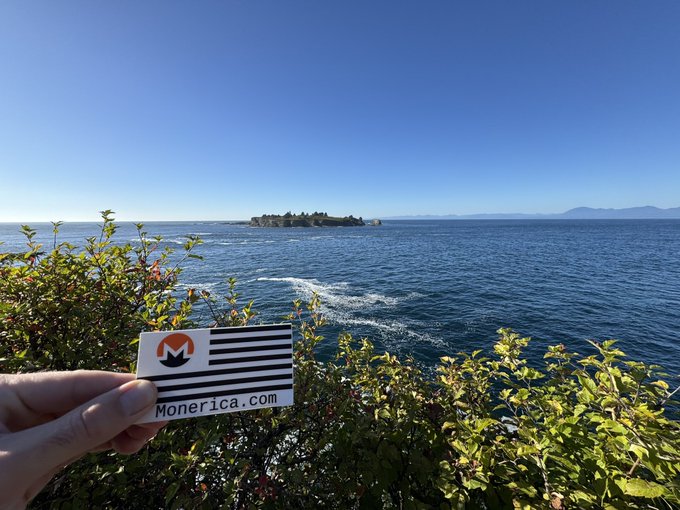 A scenic photo of a hand holding a 'Monerica.com' card with the Monero logo, overlooking a blue ocean with a forested island and mountains in the distance.