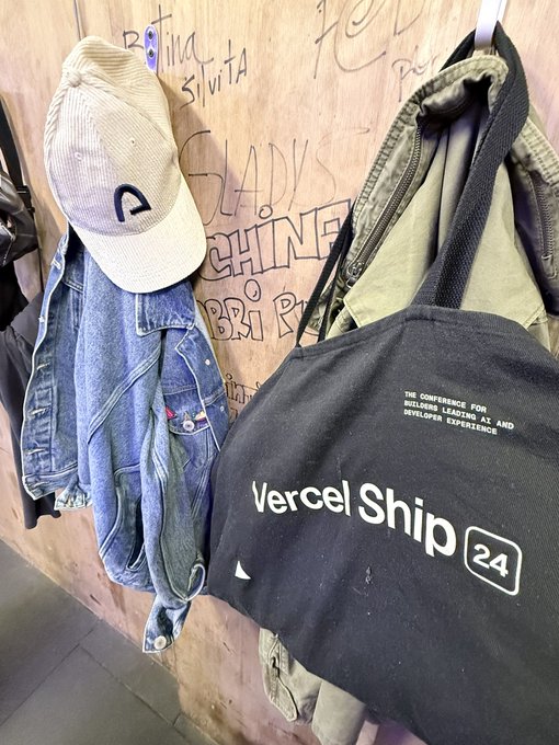 A photo of conference swag hanging on a wooden board, featuring a beige cap with a "C" logo, a denim jacket, and a black tote bag reading "Vercel Ship 24 - The Conference for Builders Leading Next AI and Developer Experience."
