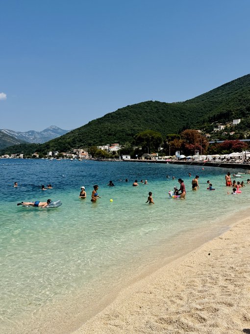 A scenic beach view with clear turquoise water where numerous people are swimming and wading, with a sandy shore in the foreground, green mountains, and a small town in the background under a blue sky.