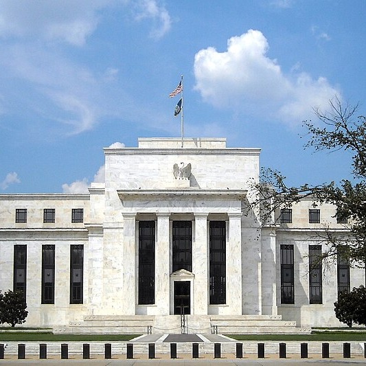 A daytime photograph of the Federal Reserve building in Washington D.C., showing its neoclassical marble facade and the flag flying above the entrance.
