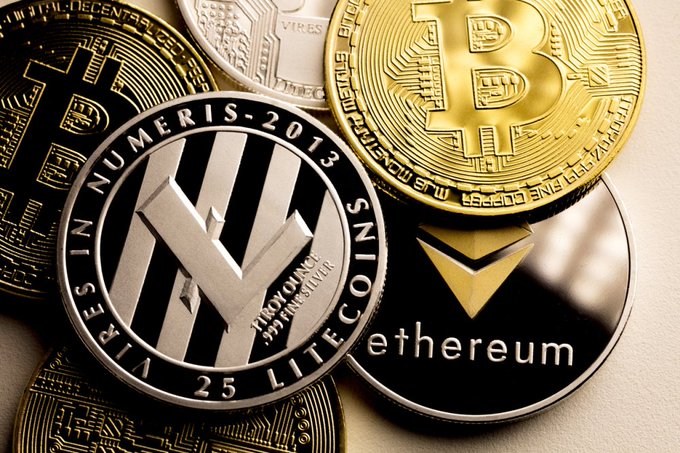 A close-up photograph of several physical cryptocurrency coins, including a silver Litecoin coin marked 'VIRES IN NUMERIS 2013', a gold Bitcoin coin, and a black and silver Ethereum coin.