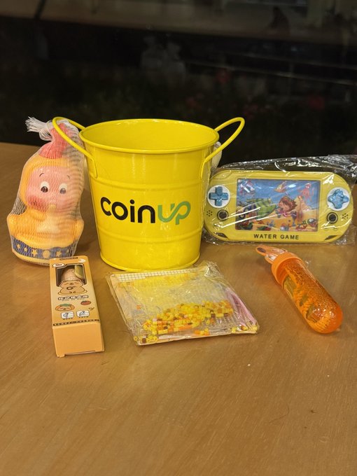 The image features a wooden table with various items, including a yellow bucket, a toy duck, a game console, and some toys. The bucket has "coinup" written on it, indicating that it's part of a promotional campaign or gift set. A small child's toy is also present on the table. The objects are arranged in an interesting manner, with the bucket being the largest item and the toys scattered around it.