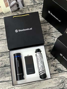 An open black gift box branded 'Stockcoin.ai' reveals a sleek cylindrical device, a black power bank, and a swirled water bottle inside.