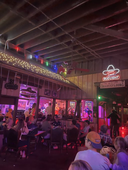 A dimly lit bar interior with colorful string lights, neon signs including 'Bud Light' and 'Jim & Eddie's Dancefloor', and patrons seated at tables watching a performance.