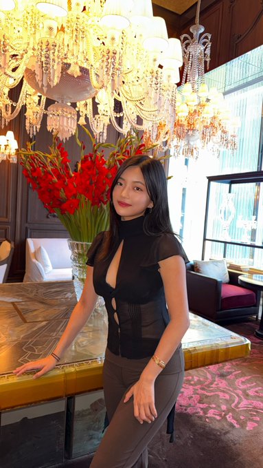 A young woman poses elegantly in a luxurious hotel lobby, leaning against a polished marble table beneath ornate crystal chandeliers with a vibrant bouquet of red gladiolus behind her. The opulent, high-end setting visually reinforces the post's confident, aspirational tone about teaching others the ropes of trading perpetual futures ("perps").
