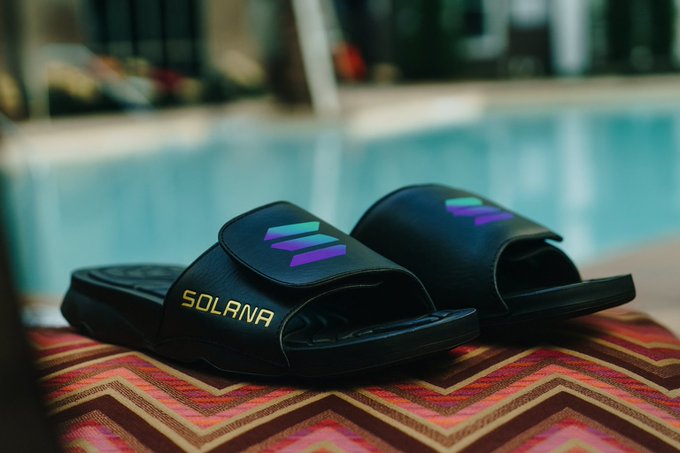 A pair of black slide sandals with the Solana logo and purple-green chevron design sit poolside on a colorful patterned surface.