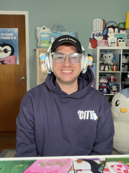 A smiling man wearing a "Knock Indoor" cap, white headphones, and a navy "CITY" hoodie sits in a penguin-themed room, with a visible poster reading "My Penguin is Me" on the door and various penguin plushies and figurines displayed on shelves behind him. The penguin-filled setting playfully contrasts with the "Good morning bull market enthusiasts" message, suggesting a lighthearted, bullish start to the day.
