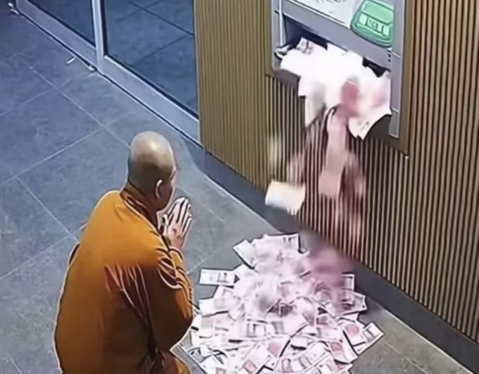 Surveillance footage shows a Buddhist monk in orange robes kneeling in prayer before an ATM that is dispensing a large pile of banknotes onto the floor.