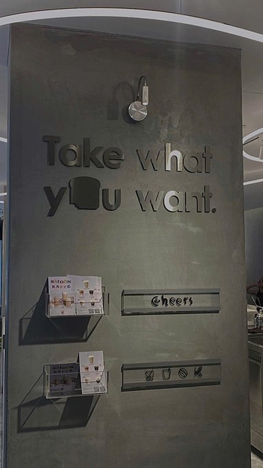 An interior wall sign reads "Take what you want" with a pot icon, accompanied by smaller "Cheers" signs and shelving displaying coffee product boxes and icons.