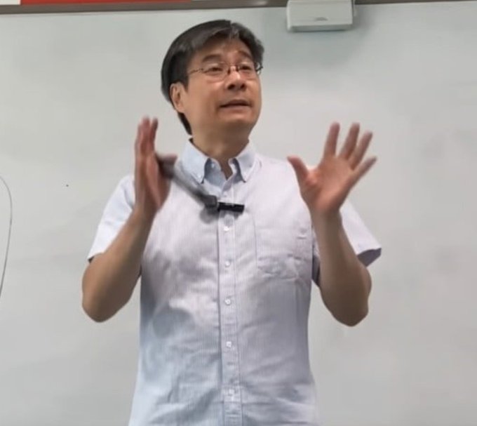 A middle-aged man wearing glasses and a light blue short-sleeved button-up shirt with a clipped microphone gestures with both hands raised while speaking in front of a whiteboard.