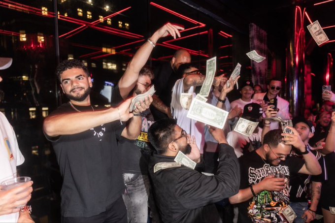 A lively nightclub scene captures a group of men throwing dollar bills in the air, with one prominent figure in a black t-shirt throwing money toward the camera.