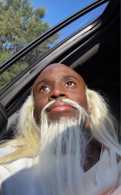 A low-angle selfie of a bald man with a long, flowing white beard and hair, sitting in a car with a sunroof open to a blue sky and trees.