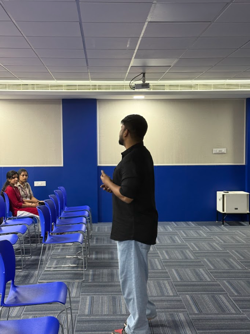A man in a black polo shirt stands in a room with blue and white walls, giving a presentation to a few seated attendees in blue chairs, with a projector mounted on the ceiling.