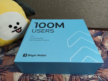 A blue box labeled '100M USERS' celebrating 'Training Crypto and Everyday Finance' sponsored by Bitget Wallet sits next to a yellow plush toy.