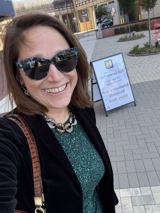 Selfie of a smiling woman wearing sunglasses, a green patterned dress, and statement necklace, standing outside an Emory location with an upside-down 'Fuji Green Egg Empanadas' sandwich board visible.