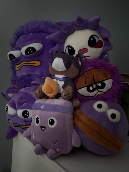 A pile of purple plush toys including duck-like characters with googly eyes, a brown dog plush with a heart tag, and a round purple character with an orange beak.