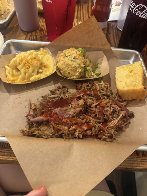 A barbecue meal tray containing pulled pork with BBQ sauce, mac and cheese, potato salad, and cornbread, served on butcher paper.