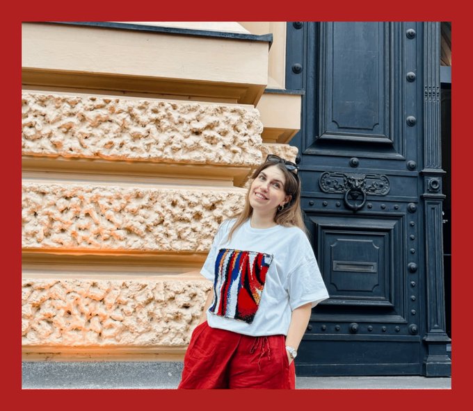 A young woman with sunglasses on her head poses in front of an ornate stone wall and a large black door, wearing a white t-shirt with a colorful patterned square design and red pants.