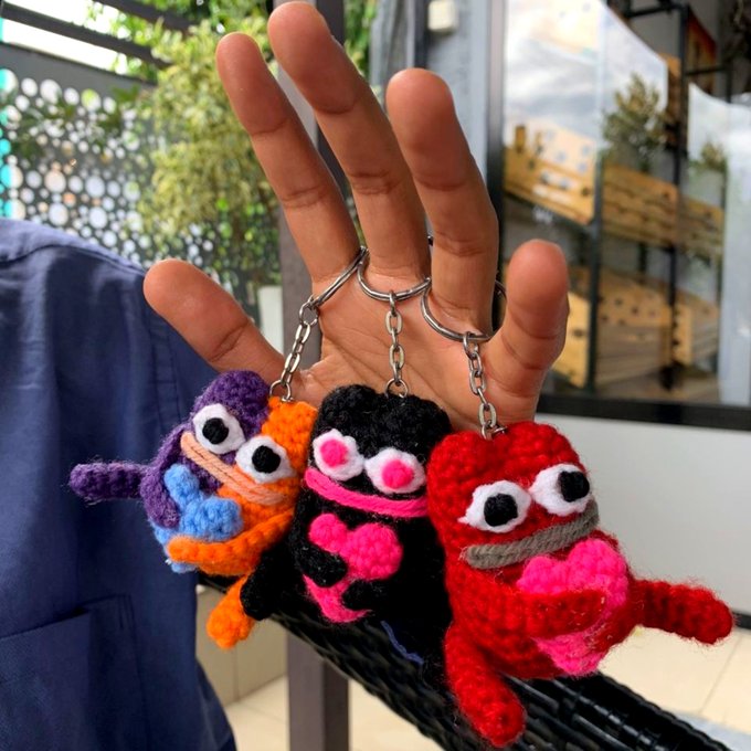 A hand holds up three crocheted monster keychains in purple/orange, black/pink, and red/pink colors.
