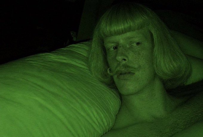A person with a blonde bob wig and mustache reclines against a pillow, bathed in green-tinted lighting. The eerie, dreamlike aesthetic suggests a luxurious or surreal scene that ties into the "GM MILLIONAIRES" theme through its stylized, wealth-evoking imagery.