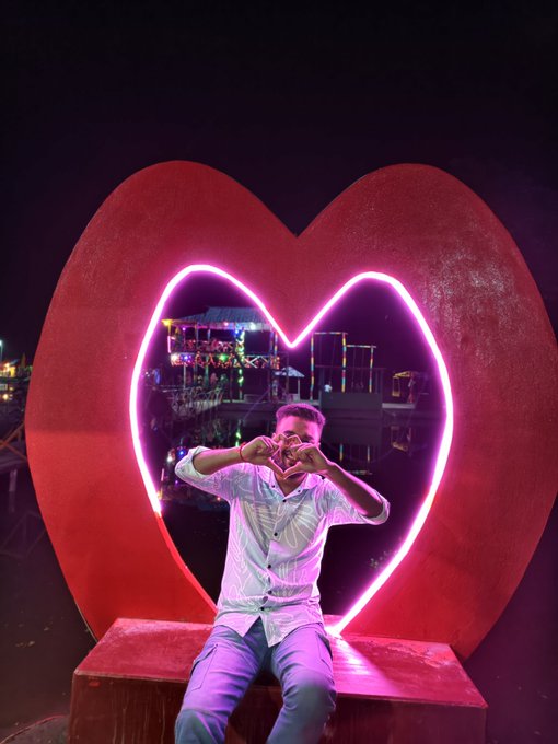 iced glass sculpture of a red heart, illuminated by pink neon lights, with a person seated on the bench and a city skyline in the background.