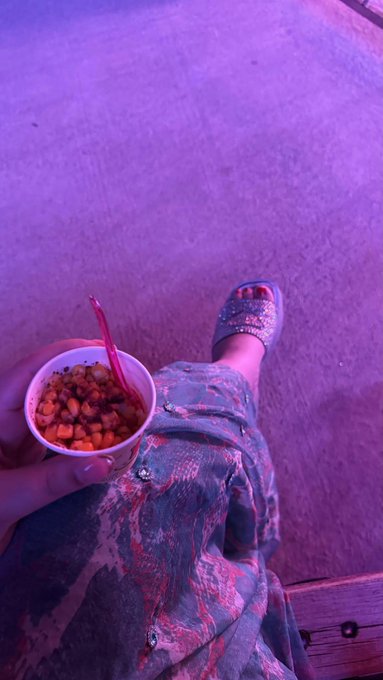 A first-person view shows a hand holding a cup of fruit cocktail with a spoon, while the other foot wears a glittery sandal, all bathed in purple lighting.