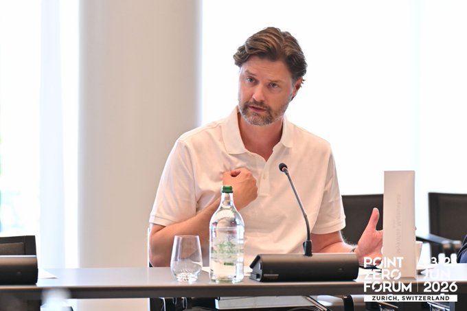 A man in a white polo shirt speaks at a panel discussion at Point Zero Forum 2026 in Zurich, Switzerland, with a water bottle and name placard on the table in front of him. The setting, a professional finance conference, aligns with the post's message about tokenization and building new financial markets.