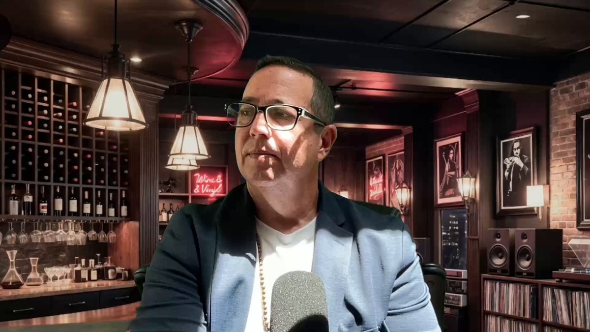 The image features a man wearing glasses, sitting at a bar with a microphone in front of him. He appears to be speaking into the microphone or recording something. The background includes several wine bottles displayed behind the man, suggesting that he might be hosting an event or discussing a topic related to wine.