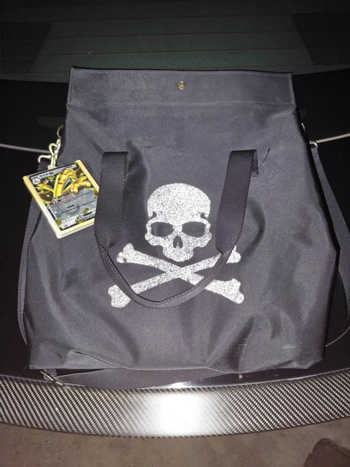 A black tote bag with a silver glitter skull and crossbones design and black straps sits on a dark surface, with a tag attached to the handle.