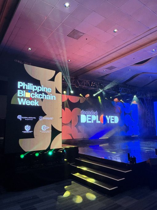 The image shows a large stage with a sign that says "Philippine Blockchain Week" and "Day 1 opened the conversation. Day 2 opens the floor." The stage has a blue and red background, and there are lights shining down on it. A few people can be seen in the scene, possibly attending the event or waiting for their turn to perform. There is also a banner that says "CoinsGeek" and "ALT+TAB", which suggests that this might be an event related to gaming and music festivals.