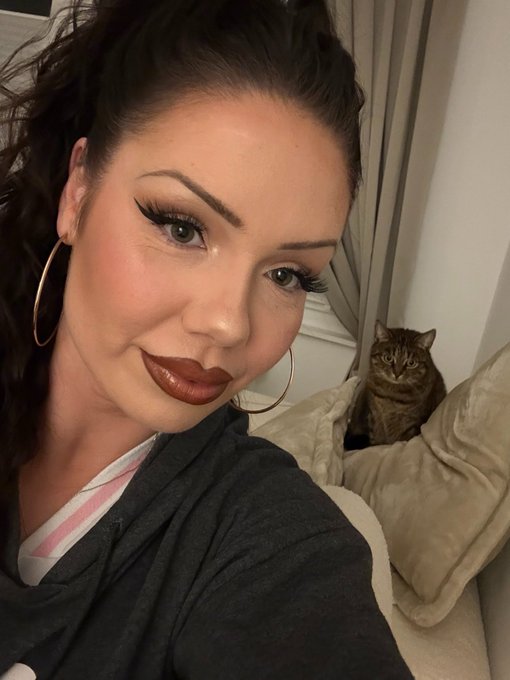 A woman with dramatic makeup poses for a selfie while a tabby cat sits on a plush bed behind her, celebrating International Cat Day with her furry companion.