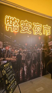 A nighttime view of the 栄安夜市 (Rongan Night Market) entrance with a glowing yellow neon sign, showing crowds of people gathered among stalls and lanterns.