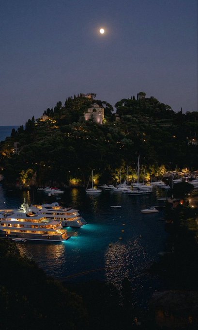 A nighttime view of a coastal harbor featuring luxury yachts illuminated in turquoise water, with a hillside village and a full moon in the sky.