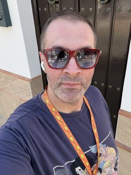 A selfie of a man wearing red-framed sunglasses and white wireless earbuds, with an orange lanyard around his neck, standing in front of a dark wooden door.