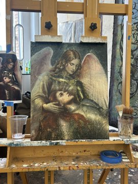 A painting of an angel embracing a human figure rests on a wooden easel in an artist's studio, surrounded by paintbrushes and supplies.