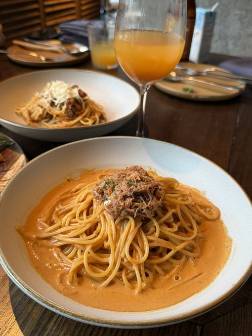 Two bowls of spaghetti on a wooden restaurant table, one with a creamy orange sauce topped with shredded meat, the other with traditional tomato sauce and cheese, alongside a glass of orange beverage.
