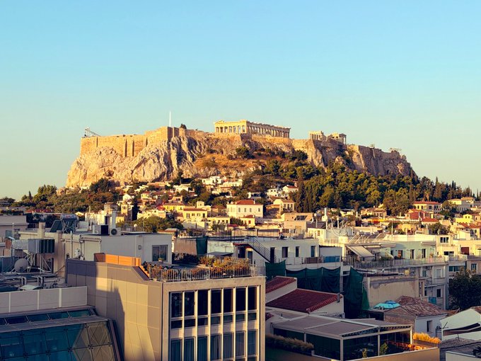 A scenic view of the Acropolis in Athens, Greece, featuring the Parthenon atop a rocky hill with the city spread below under a clear sky.