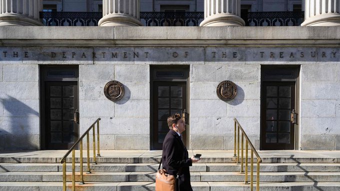 A woman walks past the entrance of the U.S. Department of the Treasury building, with its engraved name visible above the doors, symbolizing the agency's role in the new sanctions against Iranian crypto exchange BitBank for allegedly facilitating digital sanctions evasion tied to the IRGC and Strait of Hormuz shipping.
